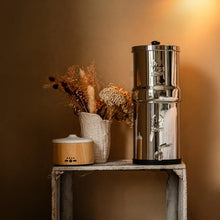 A tall stainless steel water filter pitcher with a metal spout and lid, displayed on a rustic white table next to a small wooden diffuser and a vase of dried flowers against a warm brown wall.