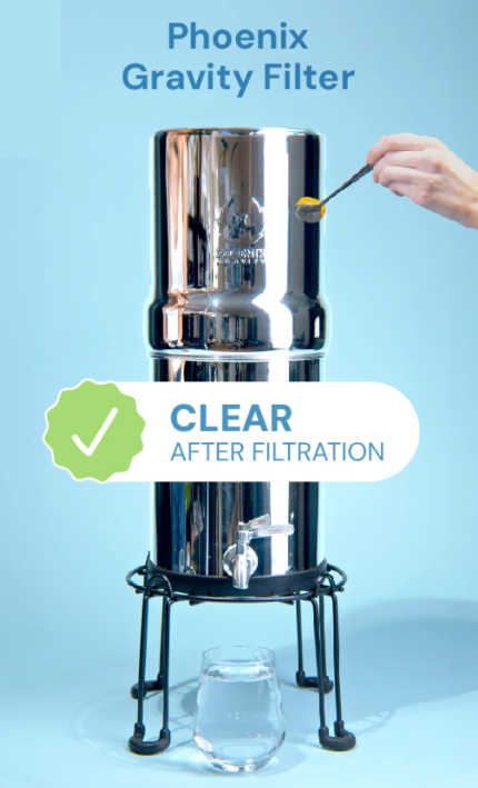A Phoenix Gravity Filter water purifier with a shiny stainless steel cylinder on a black metal stand, shown with a glass of water and a hand adding lemon. Text overlays read 'Phoenix Gravity Filter' and 'CLEAR AFTER FILTRATION' on a light blue background.