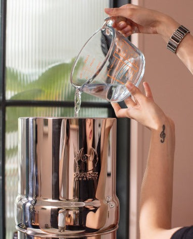 Person pouring water from a clear glass pitcher into a silver ice bucket.